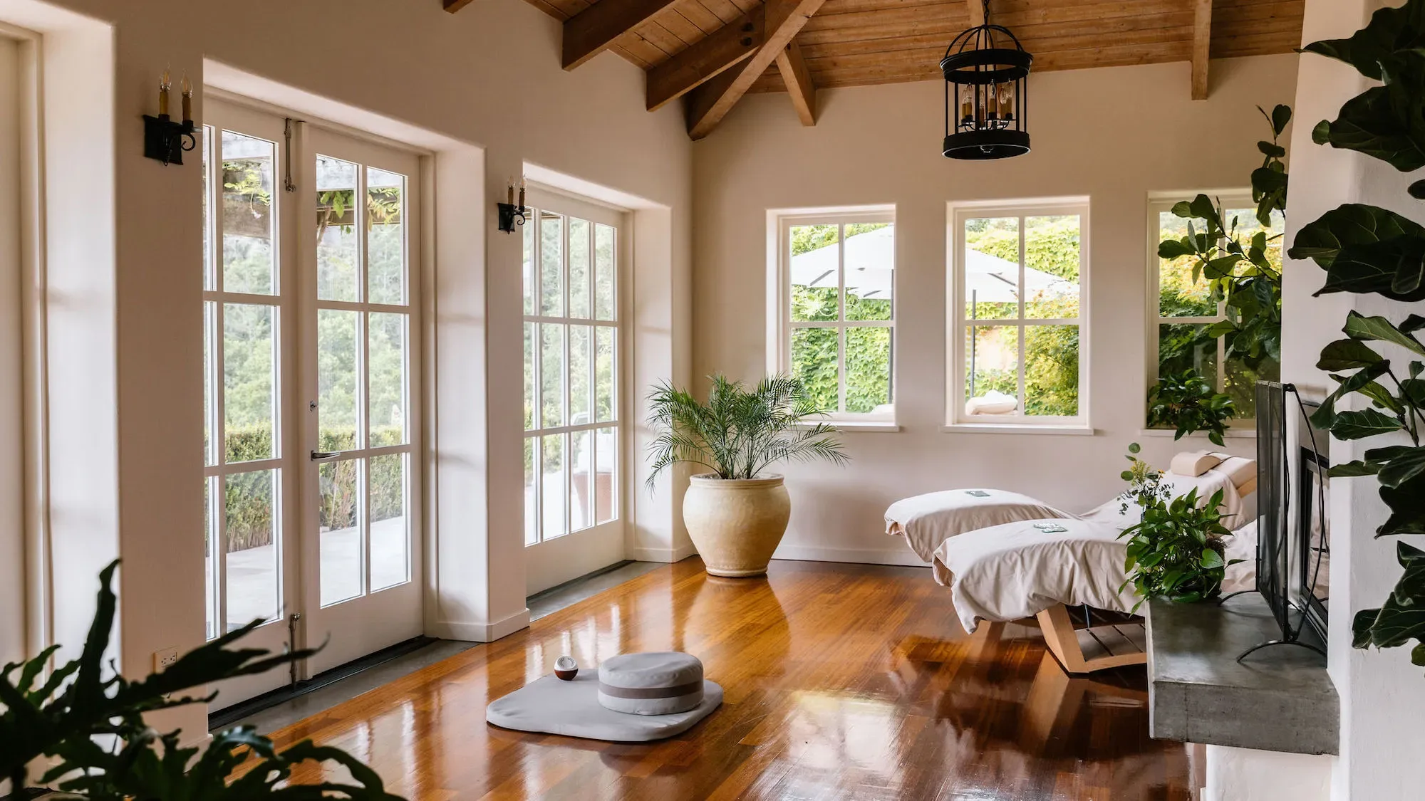 Spa room with beamed ceiling, lantern, French doors, cream beds by a fireplace, cushion, and palms, at Auberge du Soleil in Napa Valley.