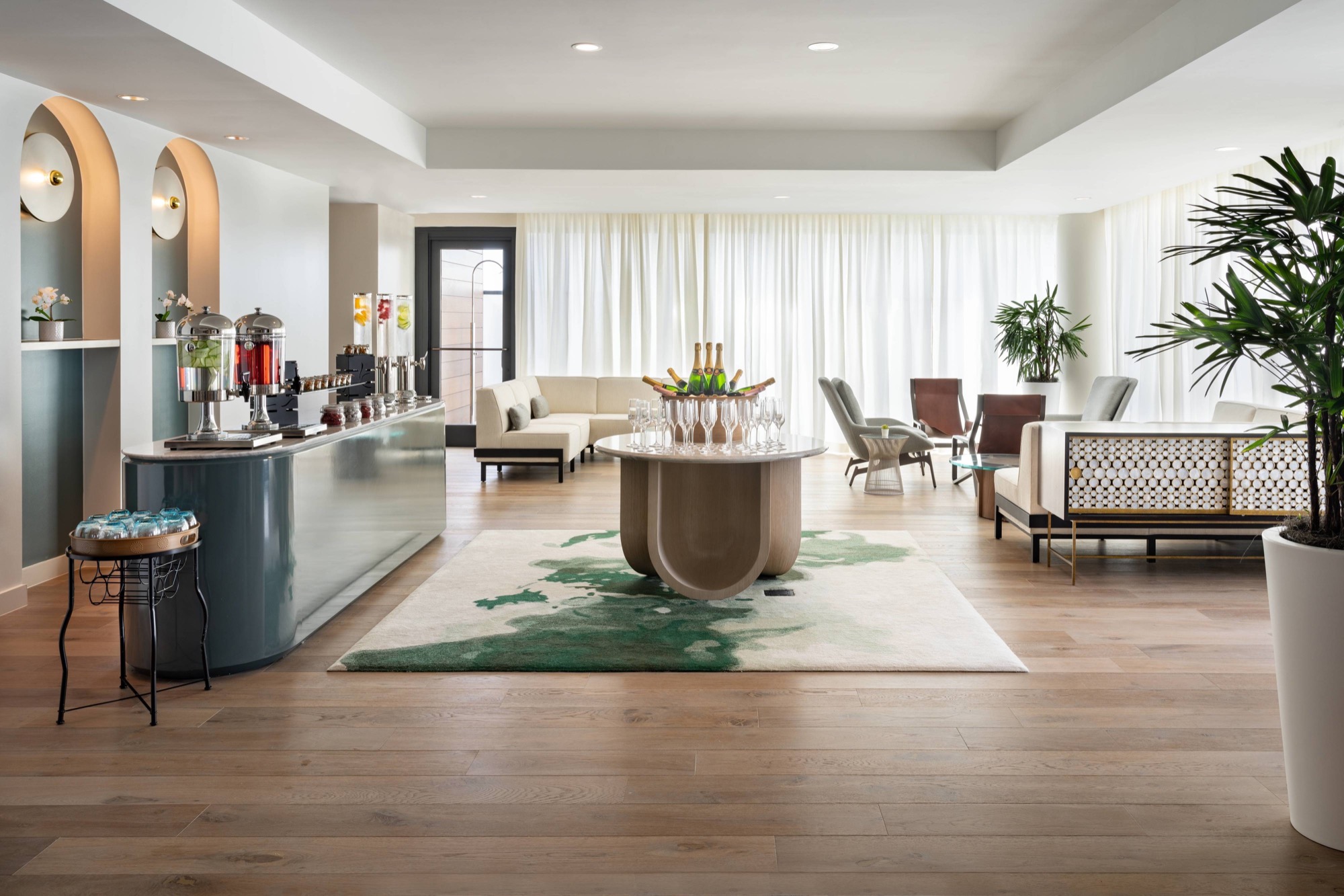 Sofas and armchairs sit around a round wooden table on a green rug, curved blue-green counter left, tall plants behind.
