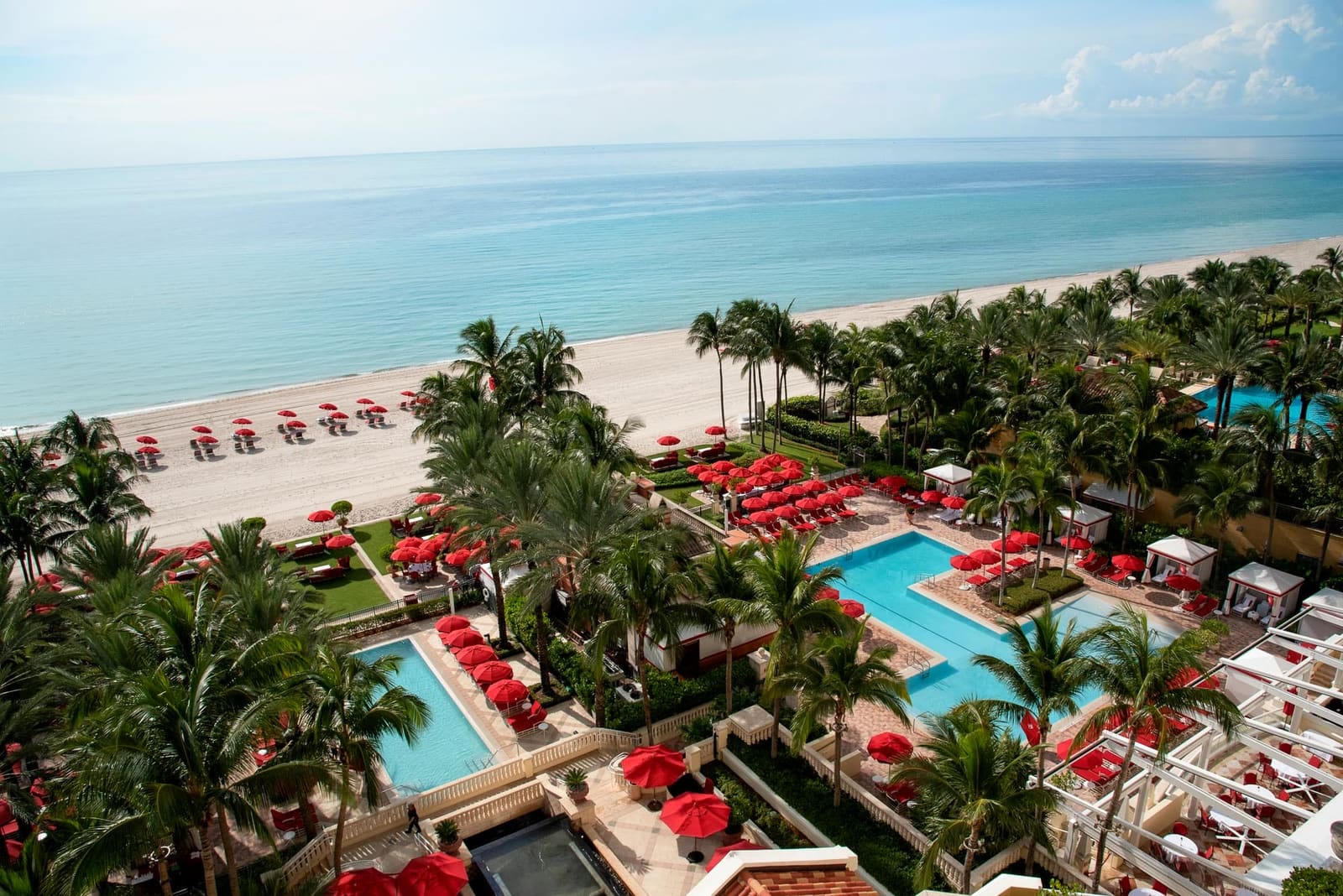Looking down on two pools ringed by palms and red umbrellas, a wide sand beach with more red umbrellas, and the turquoise Atlantic to the horizon.