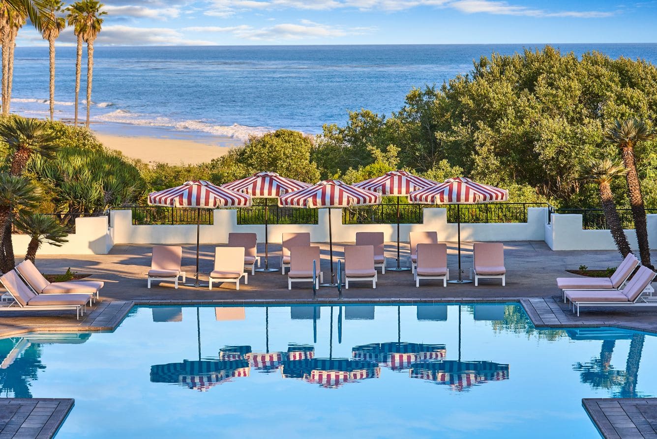 Red-and-white striped umbrellas over pink loungers on a pool terrace, reflected in the pool, with palms, a sandy beach and the Pacific behind.