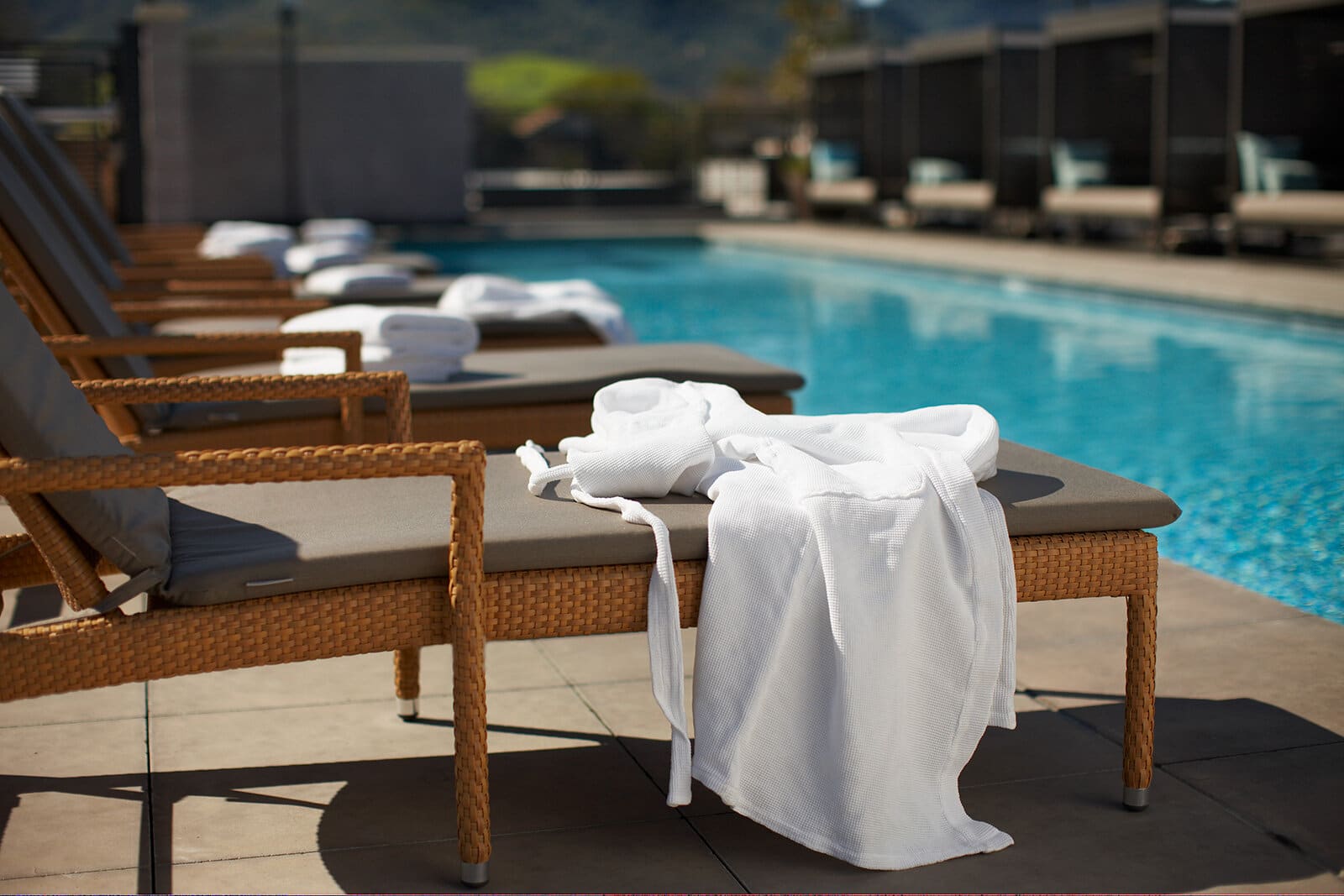 A white waffle robe draped over a wicker lounger beside a blue pool, with cabanas and hills out of focus behind.