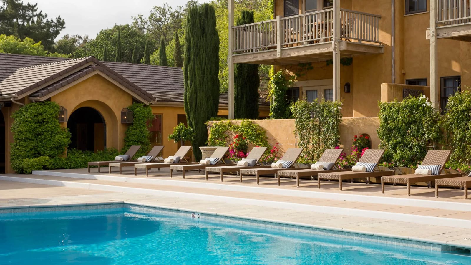 A row of wicker loungers with striped cushions along a pool deck in front of an ochre villa with balconies, cypress trees and flowering shrubs.