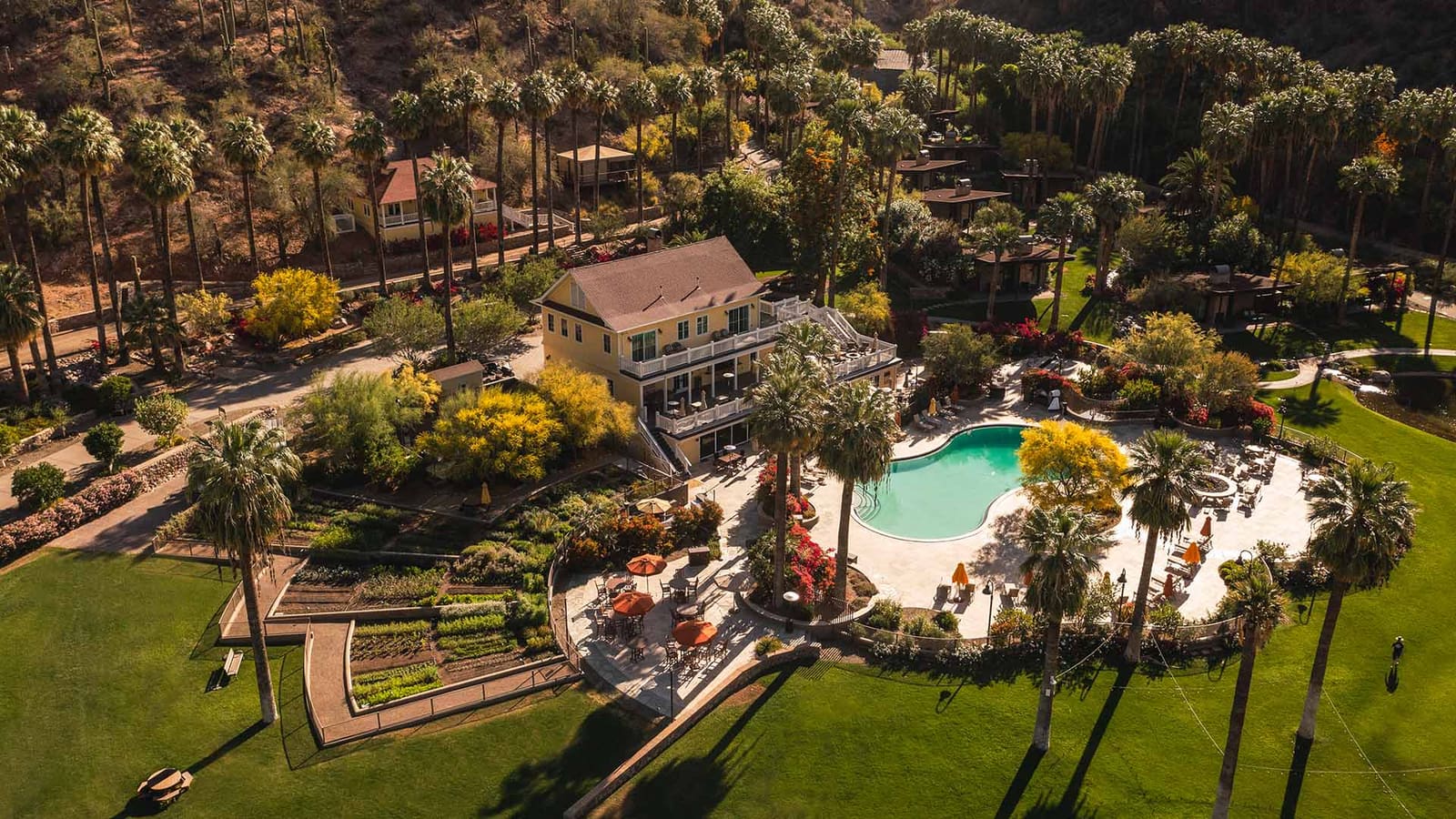An aerial view of a yellow main house beside a kidney-shaped pool, ringed by palm groves, gardens and lawn, with a desert hillside behind.