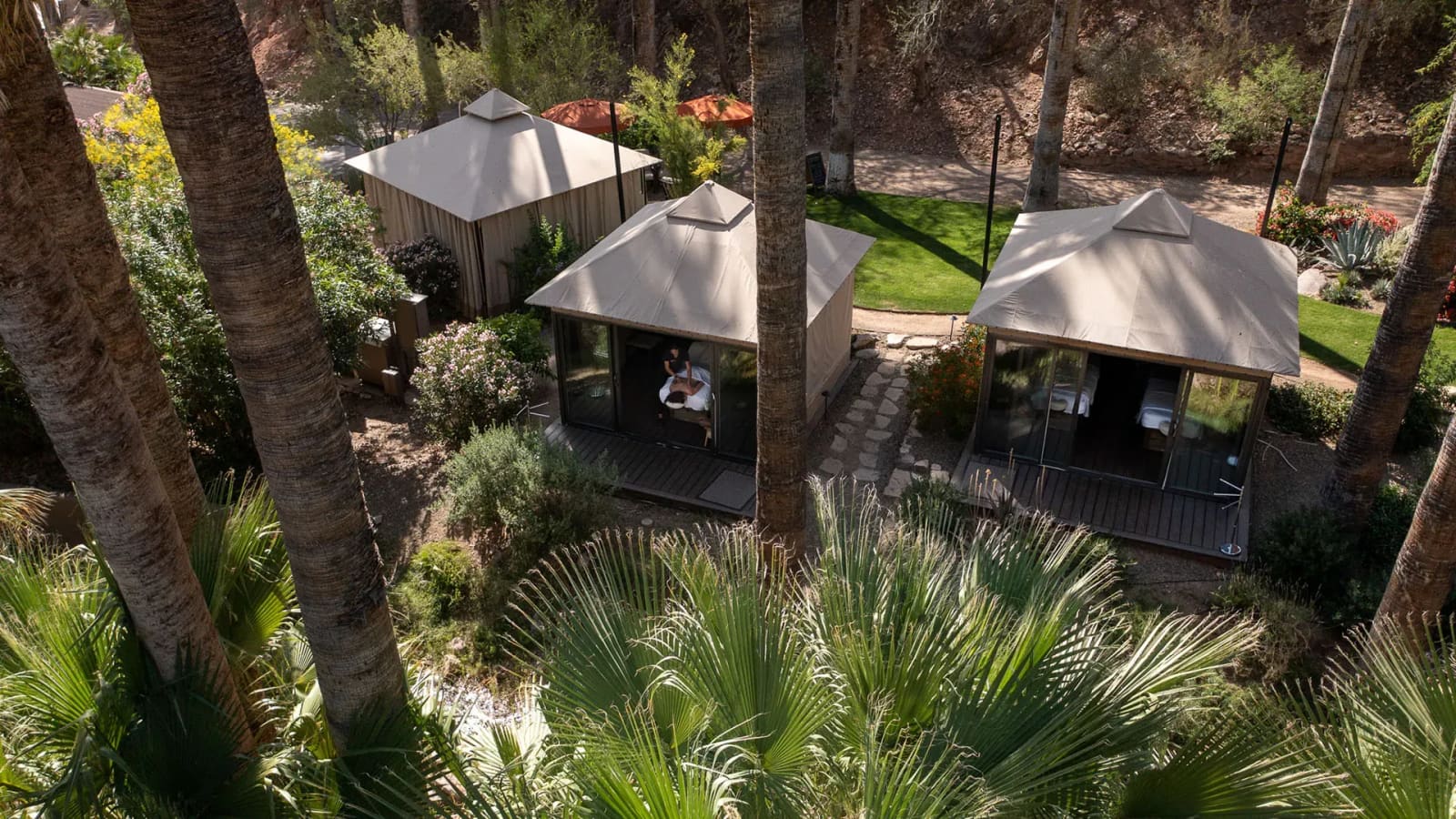 Looking down through palm fronds on canvas spa cabins among palms, a massage visible through one cabin's glass, with green lawn and a desert hillside.