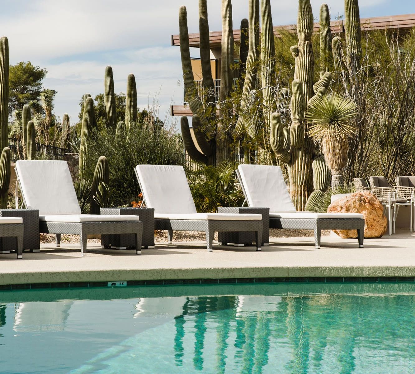 White-cushioned loungers on a pool deck in front of tall saguaro cacti, the cacti reflected in turquoise pool water.