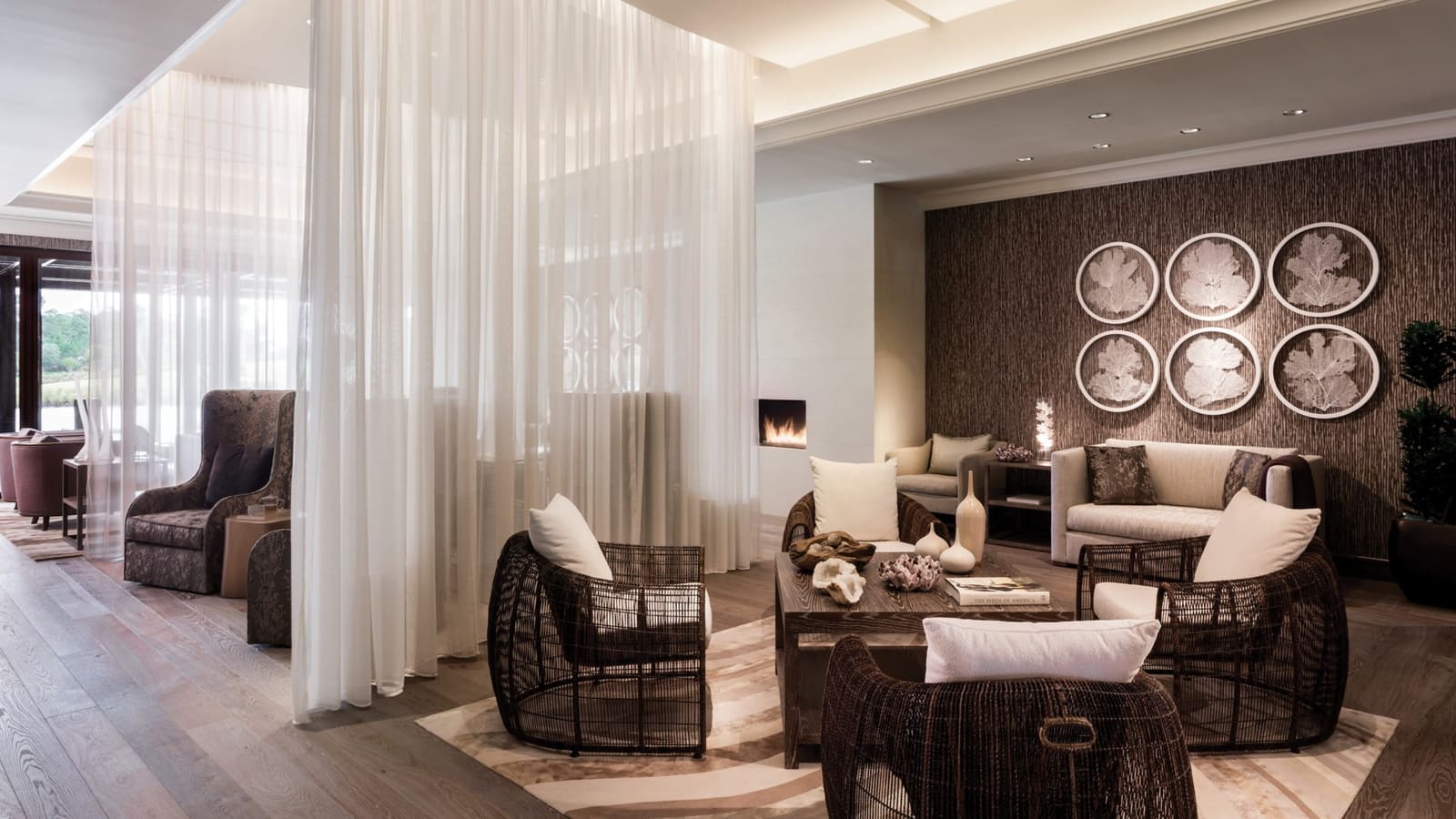 A spa relaxation lounge with sheer white curtains, wicker chairs around a low table, a fireplace and a textured wall hung with six coral-fan frames.