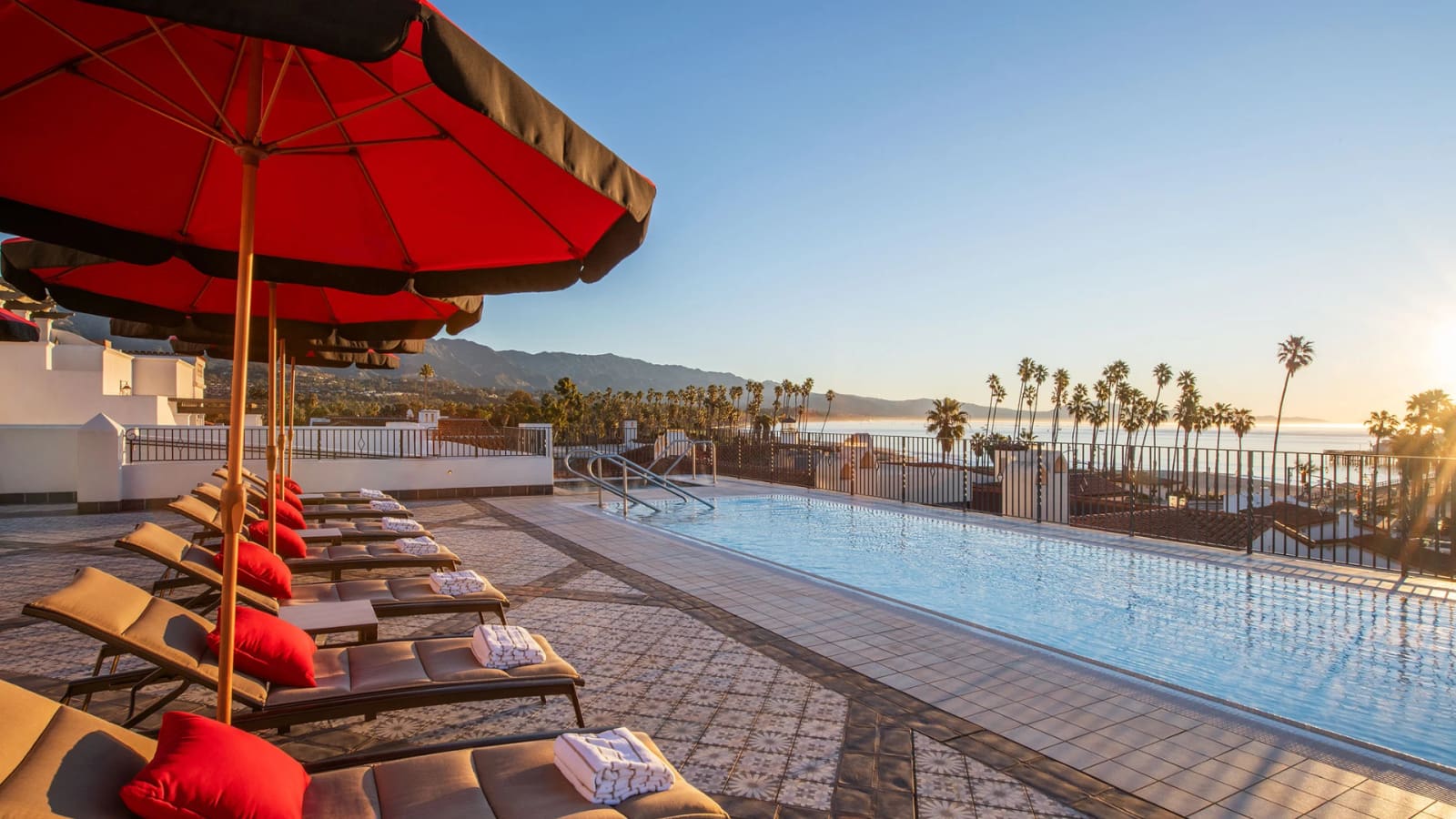 A rooftop pool at golden hour with red umbrellas and loungers along one side, palms, the ocean and mountains beyond.