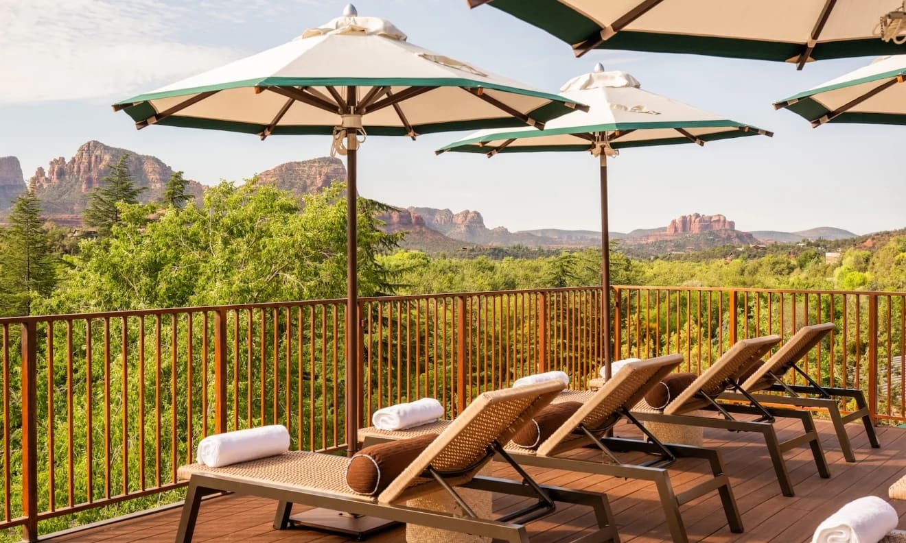 Wooden loungers with rolled white towels under green-trimmed umbrellas on a railed deck, looking over a green tree canopy to the red rocks of Sedona.