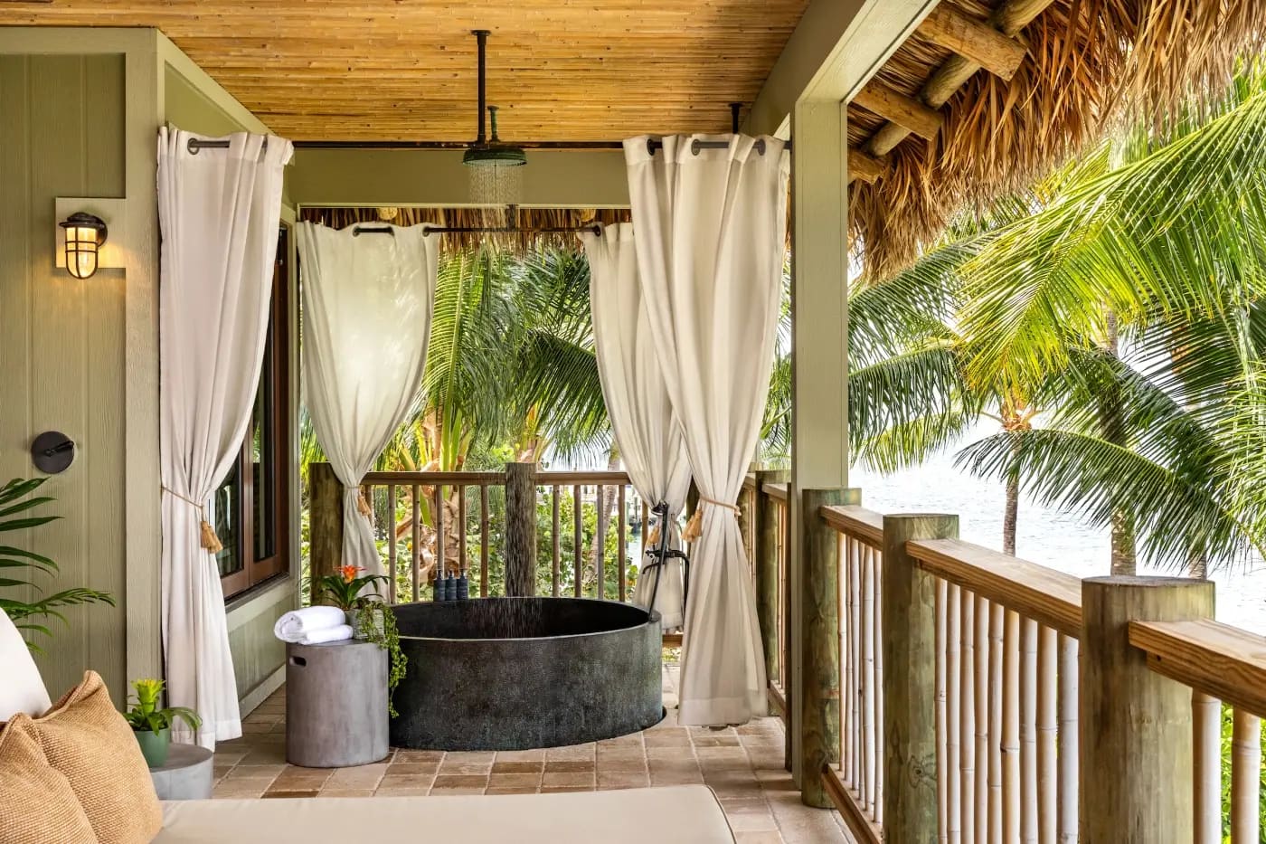 A dark stone soaking tub under a rain shower on a covered veranda, framed by white curtains and thatched eaves, with palms and water beyond the rail.