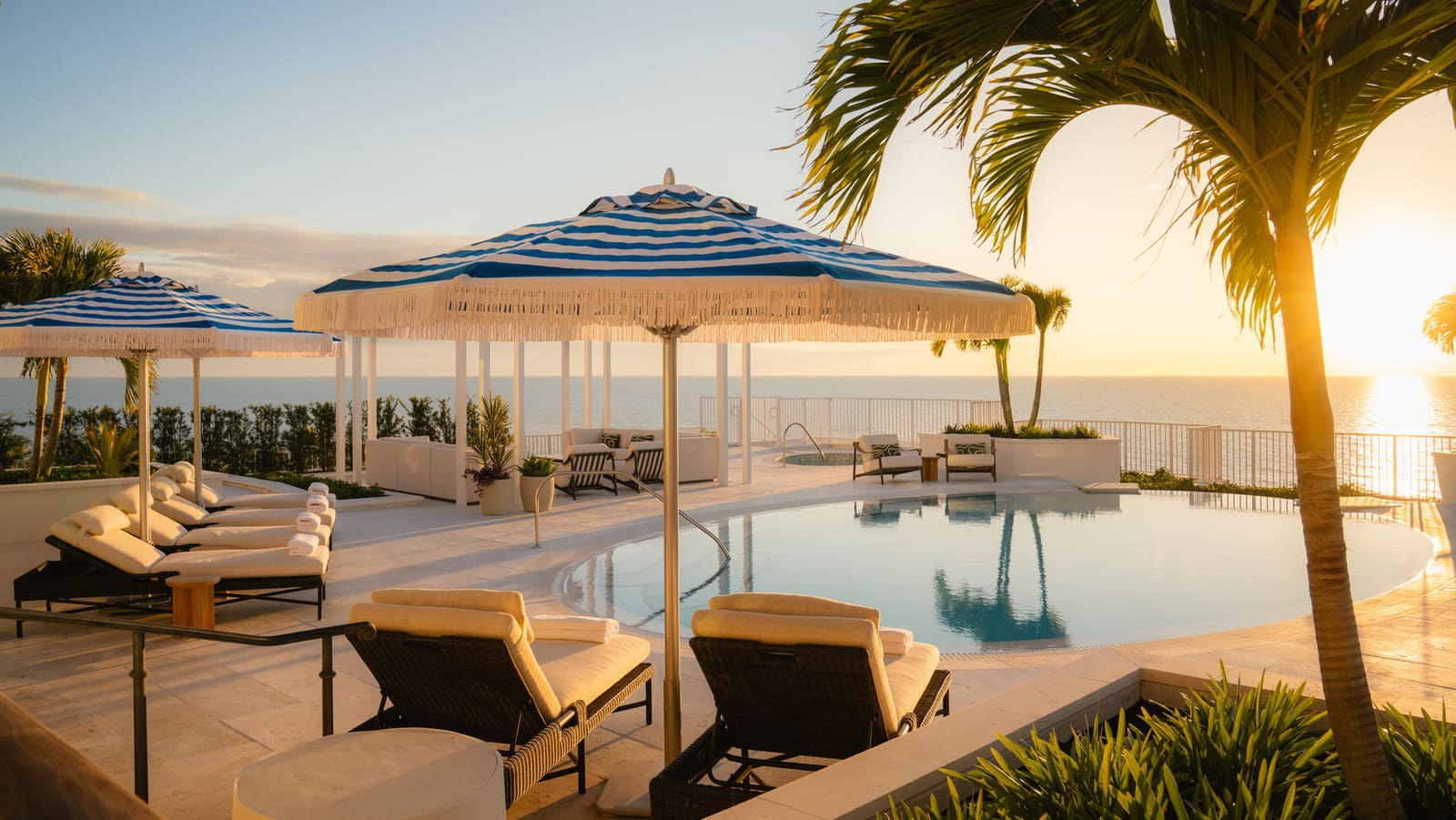 A rooftop infinity pool at sunset, blue-striped fringed umbrellas over cream loungers, a palm and the sun low over the Gulf.