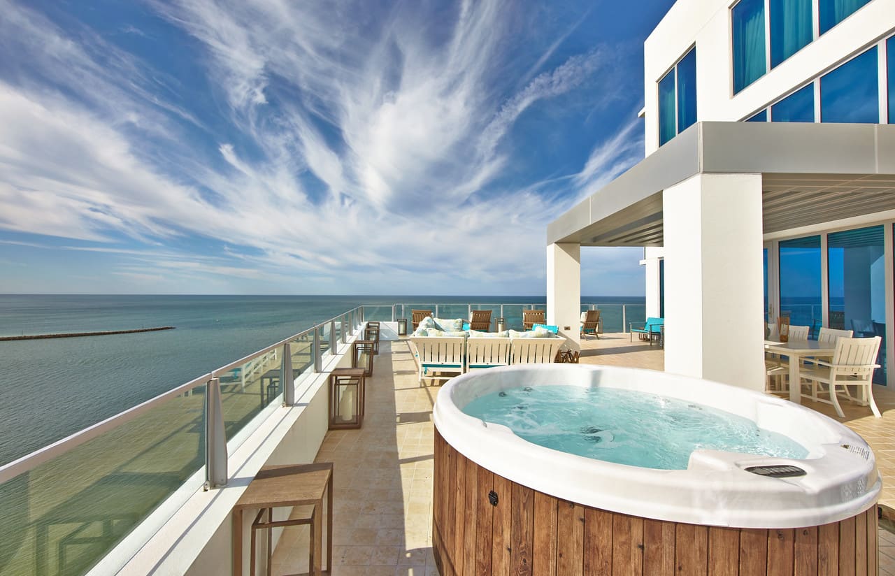 A wood-clad hot tub on a rooftop terrace with loungers along a glass rail, the Gulf horizon and a sky of wispy cloud.