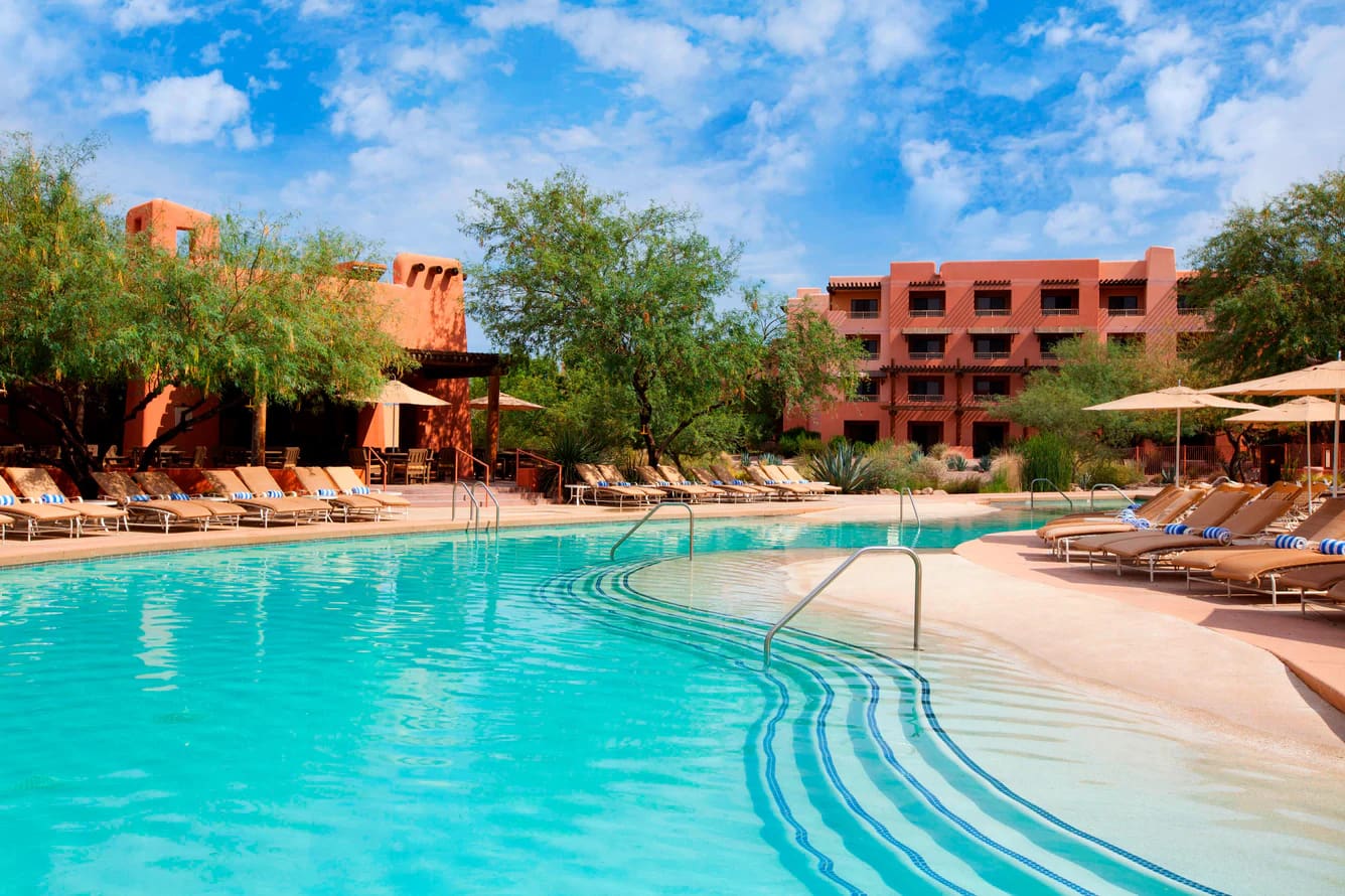 A turquoise pool with a curving zero-entry edge and handrails, loungers along the deck, and terracotta pueblo-style buildings among trees under a blue sky.