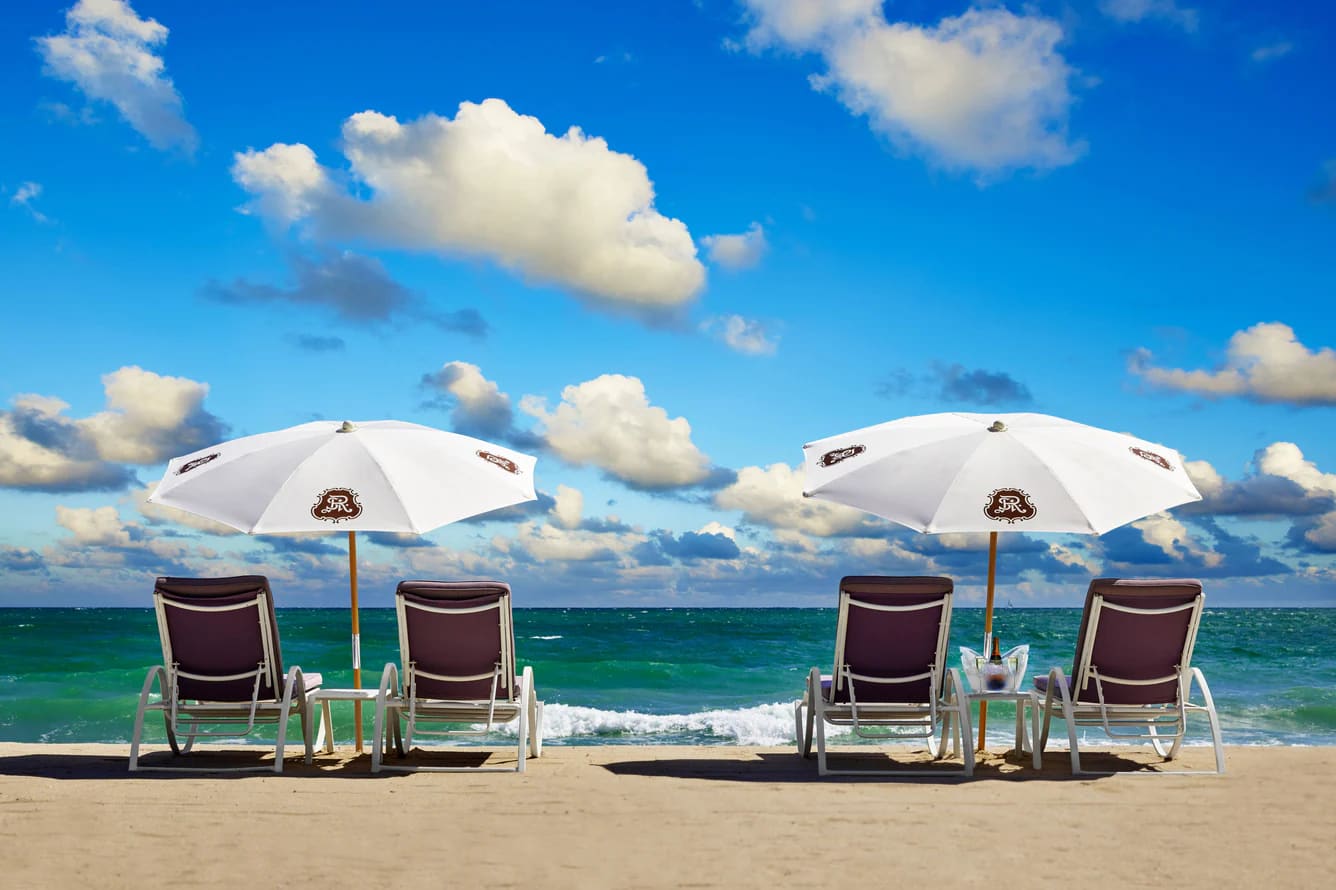 Two white branded umbrellas over four loungers on the sand, a turquoise sea and a sky of big white clouds behind.