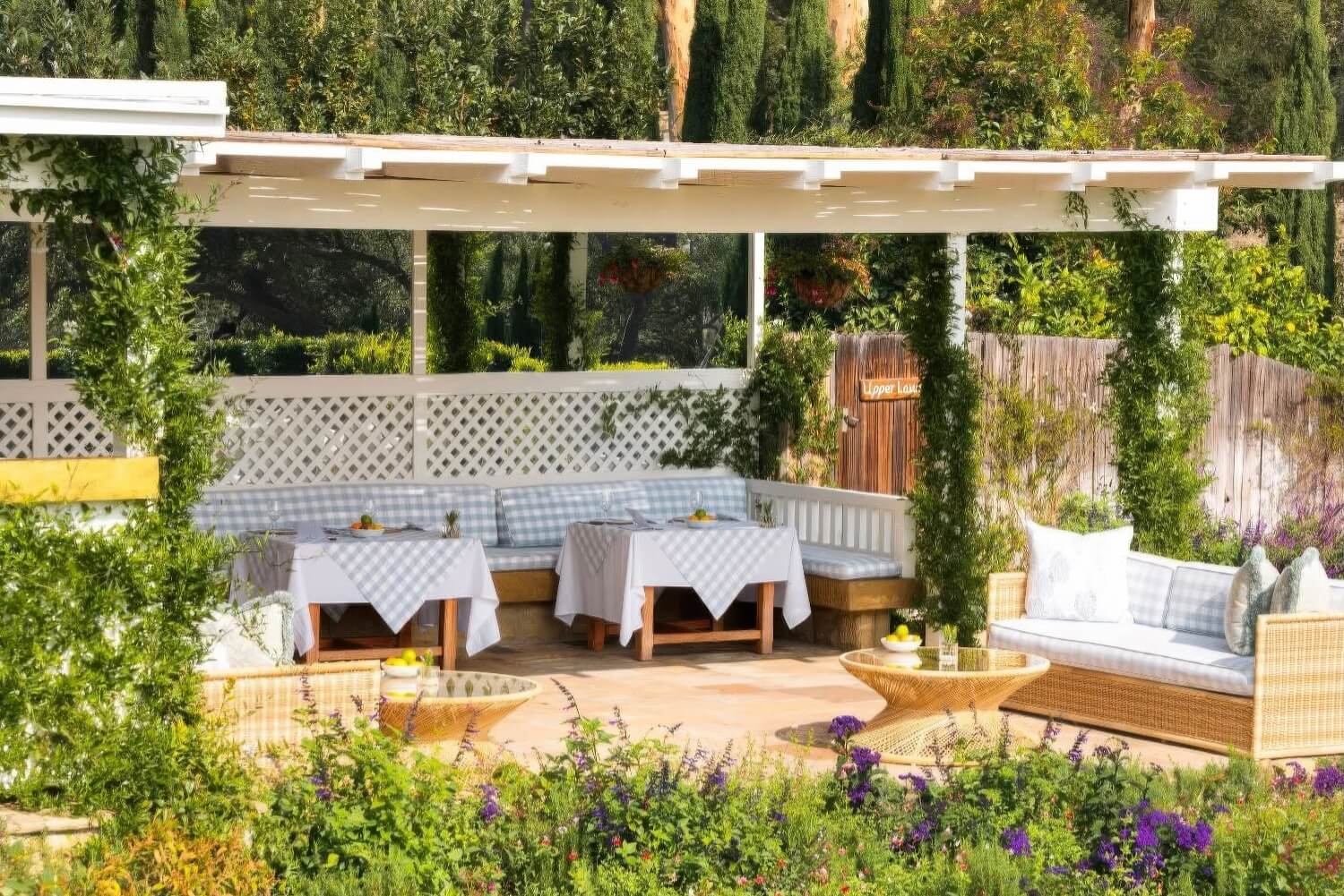 Garden dining terrace with gingham-topped tables and a wicker sofa amid climbing greenery at San Ysidro Ranch, Montecito