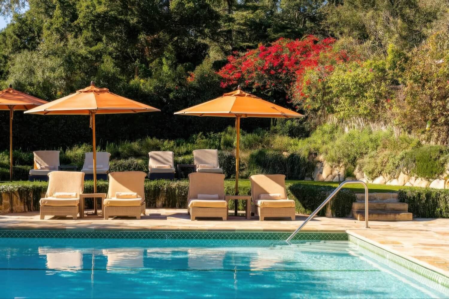 Swimming pool lined with tan loungers and orange umbrellas beside red bougainvillea at San Ysidro Ranch, Montecito