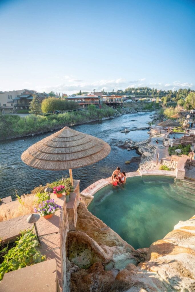 A family soaking in a riverside terrace pool under a thatched umbrella at The Springs Resort, Pagosa Springs, Colorado
