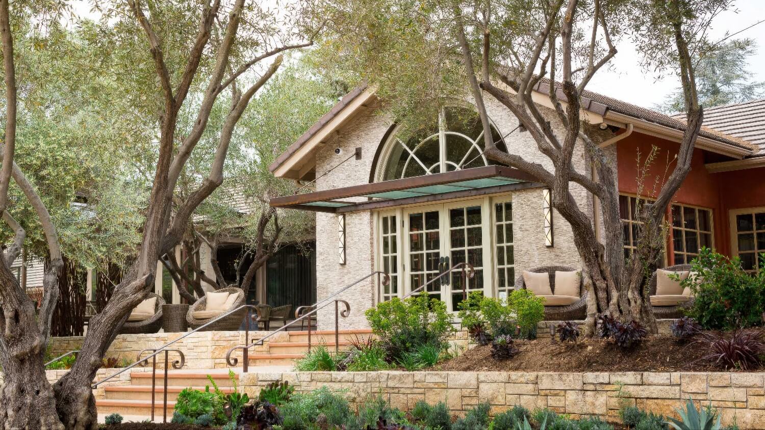 Stone lodge entrance with arched window, wicker chairs and olive trees at Bernardus Lodge and Spa, Carmel Valley