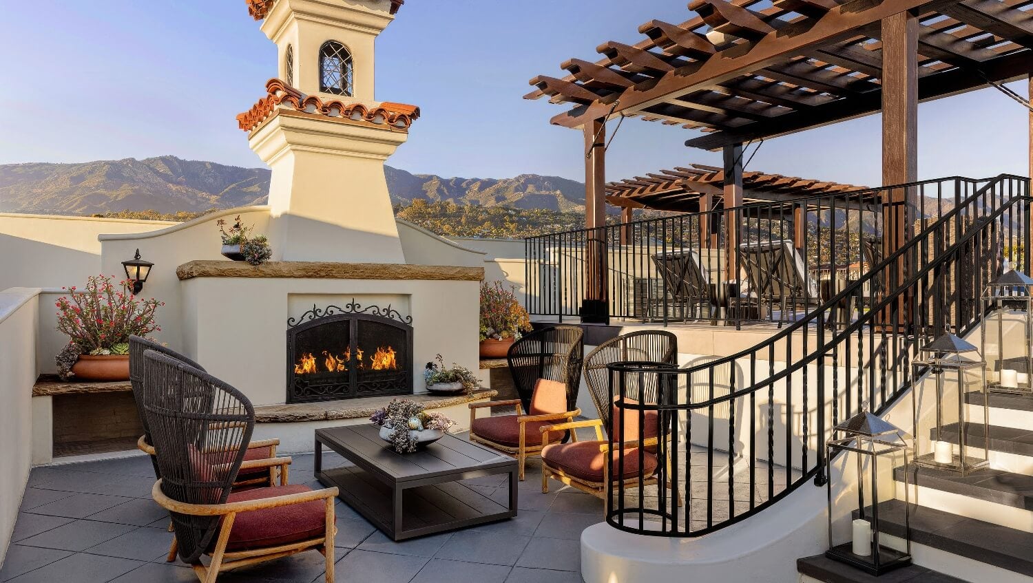 Rooftop fire terrace with woven lounge chairs beside an outdoor fireplace and tiled bell tower, mountains behind, at Kimpton Canary Hotel, Santa Barbara.