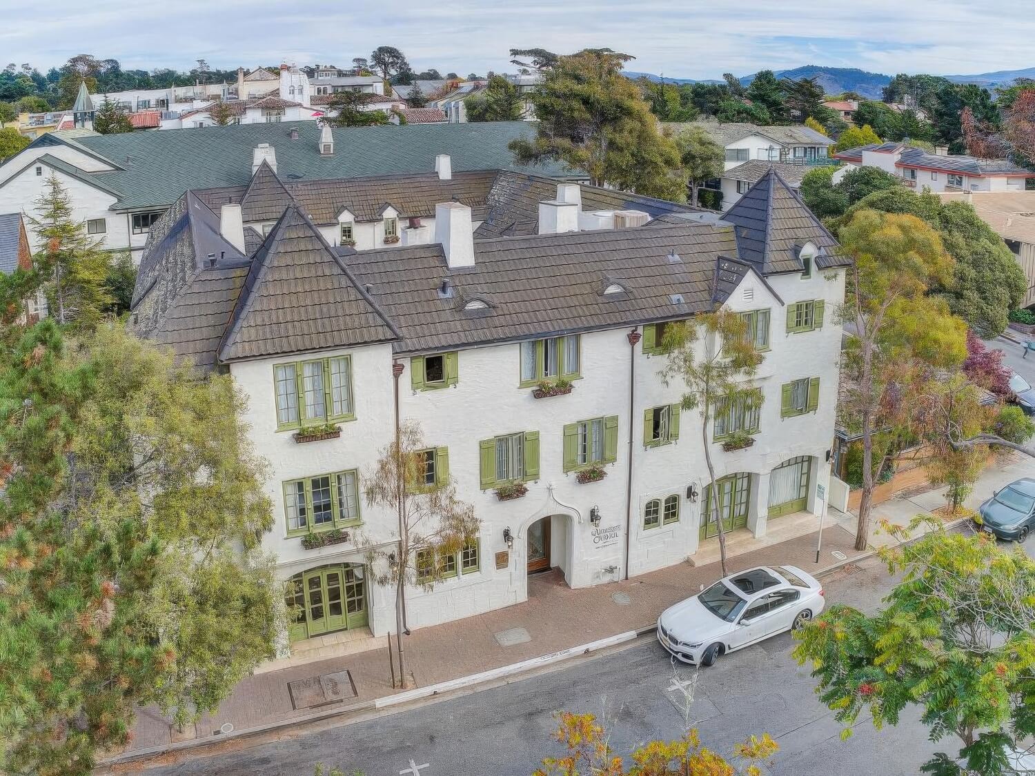 Aerial view of L'Auberge Carmel's white stucco, green-shuttered 1929 building on Monte Verde Street, with Carmel village rooftops and hills behind.