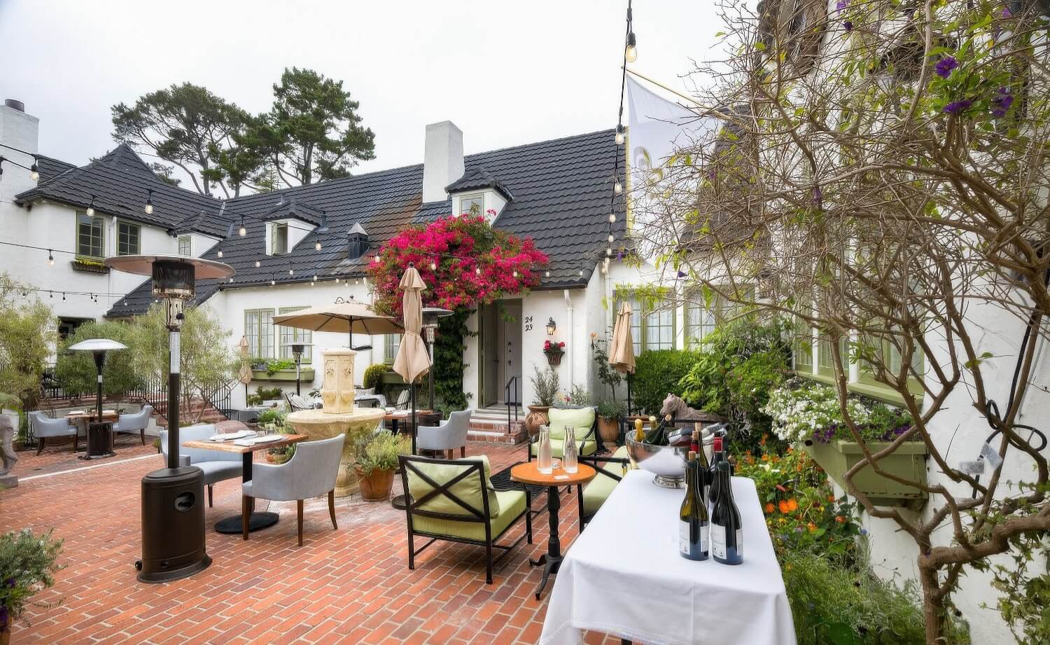 Brick courtyard set for outdoor dining with patio heaters, a wine-service table, and pink bougainvillea over a guest-room door at L'Auberge Carmel, Carmel-by-the-Sea.