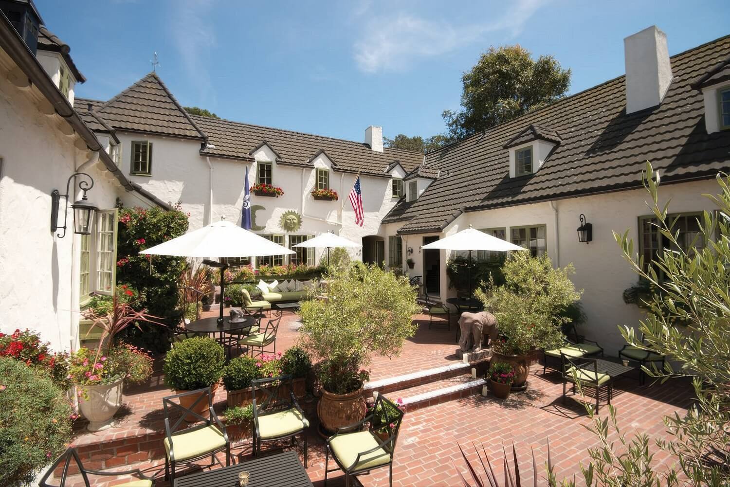 Sunlit brick courtyard with white umbrellas, potted olive trees, and sun-and-moon emblems on the white stucco walls at L'Auberge Carmel, Carmel-by-the-Sea.
