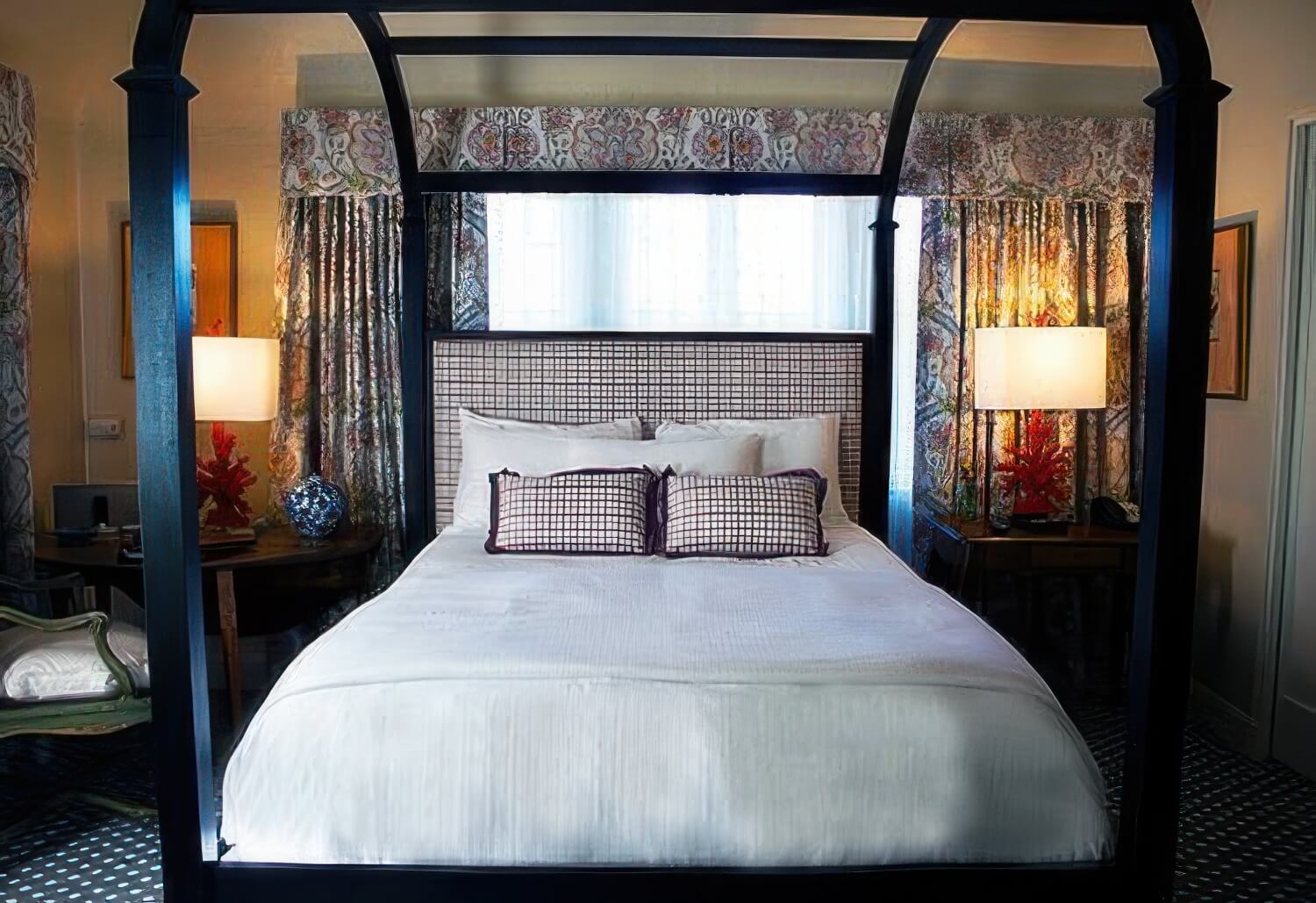 Black four-poster canopy queen bed with white linens, patterned drapes, and coral-red lamps in a Deluxe Courtyard Queen room at L'Auberge Carmel, Carmel-by-the-Sea.