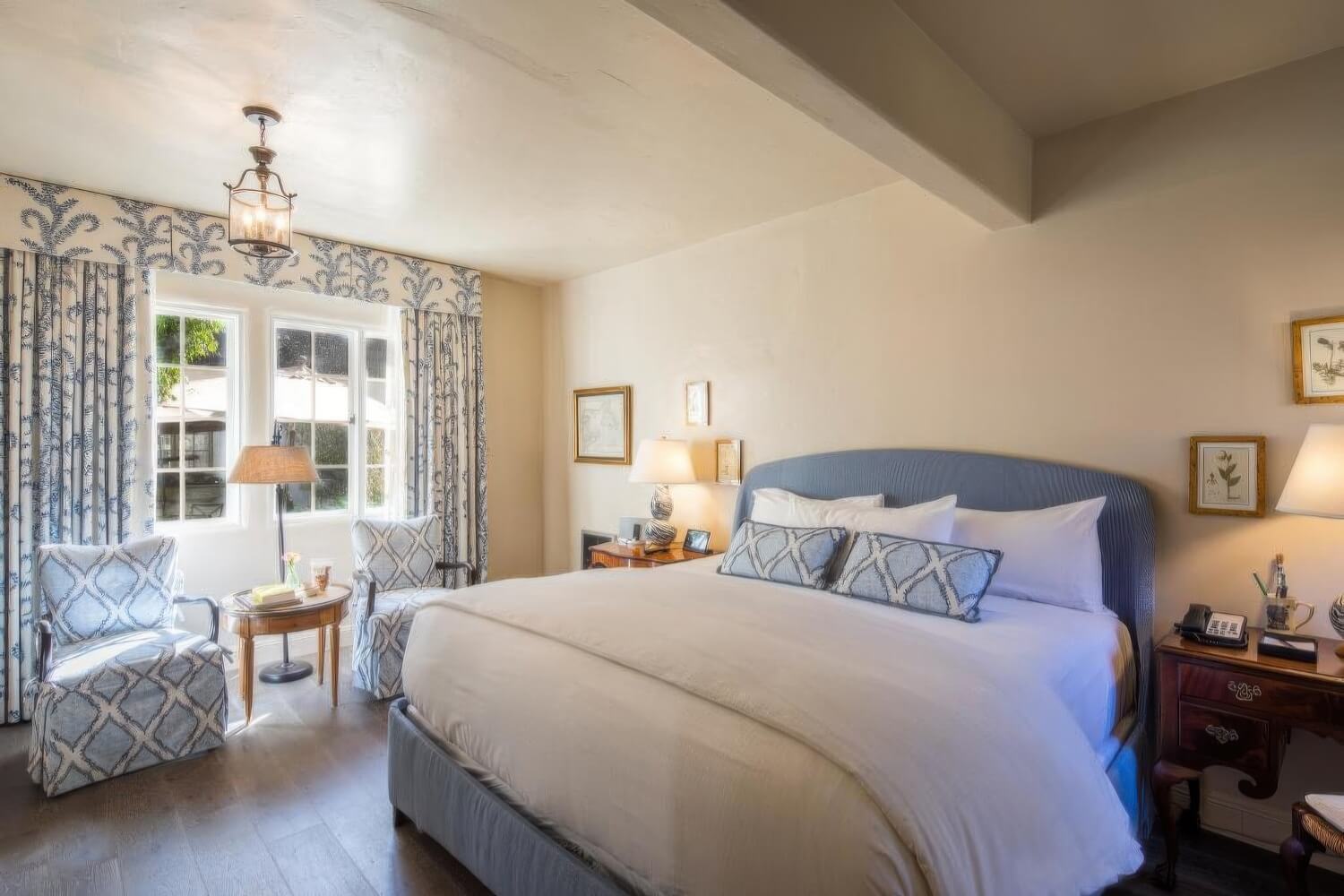 King bed with a blue upholstered headboard, blue-and-white fern-print drapes, and two patterned armchairs beside a window onto the courtyard umbrellas at L'Auberge Carmel, Carmel-by-the-Sea.