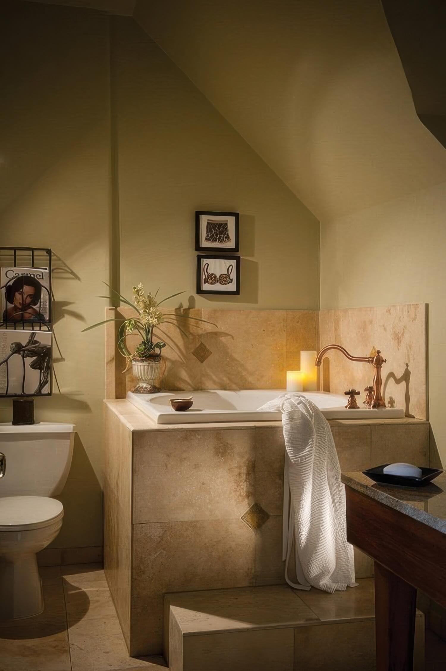 Deep tiled step-up soaking tub with a copper faucet and a lit candle beneath a sloped attic ceiling in a guest bathroom at L'Auberge Carmel, Carmel-by-the-Sea.