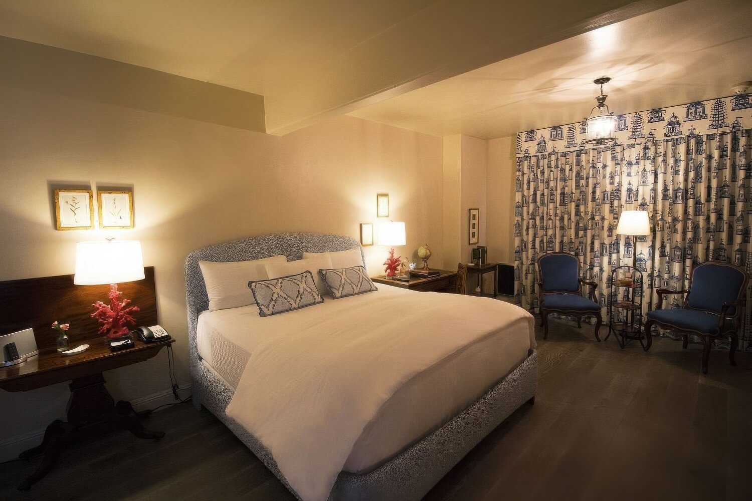 King bed with a speckled upholstered headboard, blue-and-white pagoda-print drapes, and two blue armchairs in a Standard Courtyard King room at L'Auberge Carmel, Carmel-by-the-Sea.