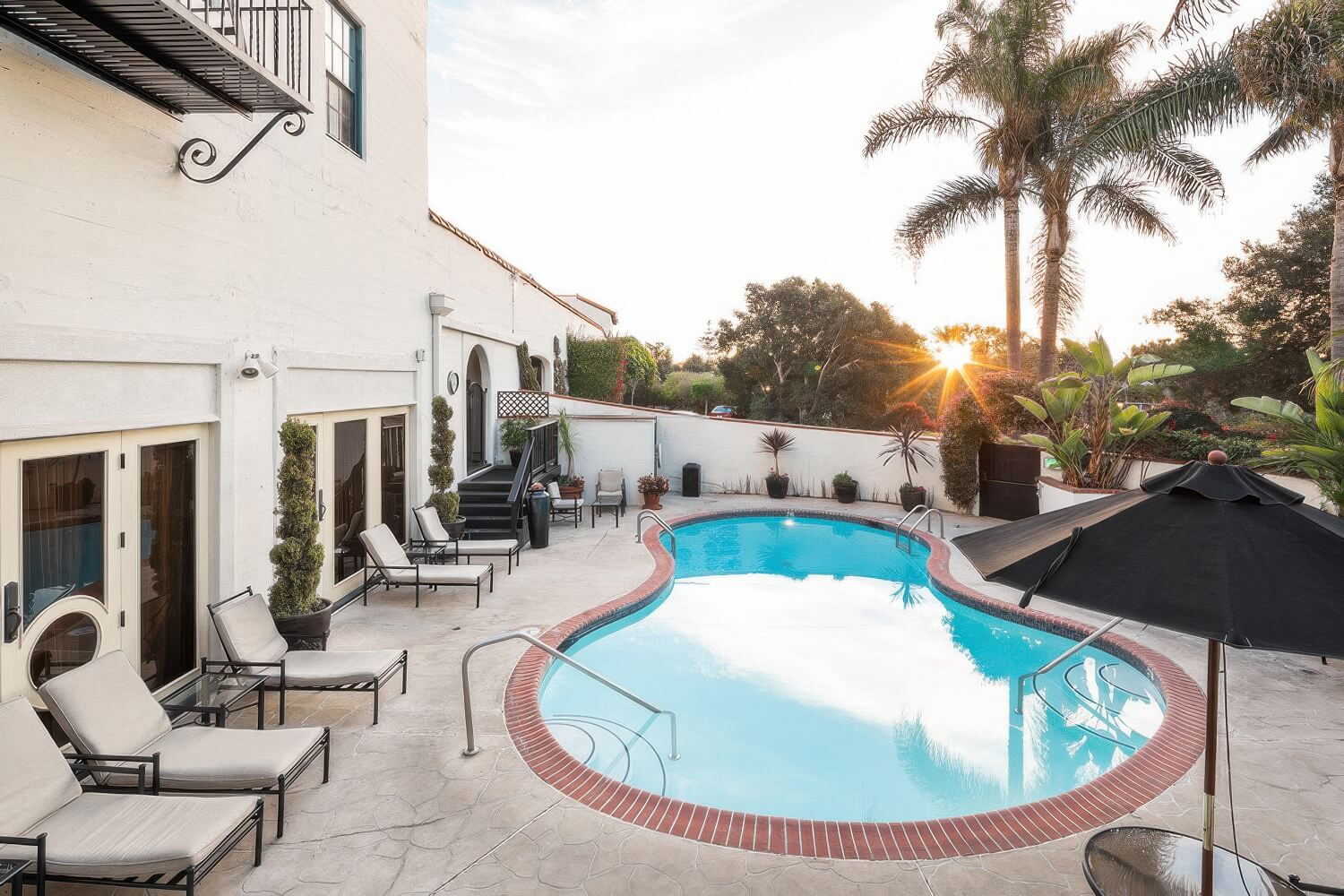 Small kidney-shaped heated pool edged in brick, with cushioned loungers and a black umbrella at sunset, at the Montecito Inn, Montecito